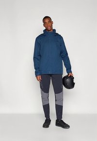 Tamsiai mėlyna vandeniui atspari striukė su gobtuvu, derinama su pilkomis ir juodomis kelnėmis, turinčiomis tekstūruotas detales. Modelis laiko juodą šalmą.