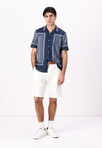 Navy short-sleeve shirt with white geometric patterns, paired with white shorts and white sneakers. Brown belt accentuates the look.