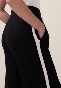 Des pantalons en tricot noirs avec des poches latérales, une taille haute et une rayure blanche contrastante le long de la couture extérieure. Texture lisse et coupe ajustée.