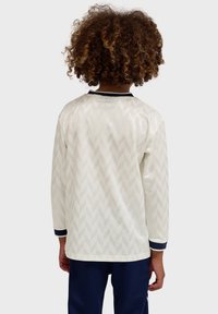 Enfant aux cheveux bouclés portant une chemise blanche à manches longues avec un motif chevron subtil et un pantalon bleu marine, vu de dos.