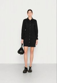 Vestido camisola de denim preto com frente de botões, dois bolsos no peito, mangas longas e corte reto. Combinado com sapatos pretos de plataforma e uma bolsa.