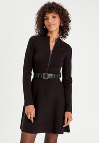 Black knit dress with long sleeves, zippered front, and a fitted waist accentuated by a wide black belt with circular cutouts.