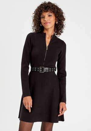 Black knit dress with long sleeves, zippered front, and a fitted waist accentuated by a wide black belt with circular cutouts.