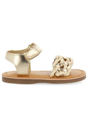 Sandal de cuero metálico dorado con una tira de dedo trenzada, correa del tobillo ajustable y una suela de goma marrón. Diseño plano apto para clima cálido.
