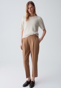 Cremfarbener, kurzärmeliger Pullover aus weichem Material, kombiniert mit braunen, hochgeschnittenen, plissierten Hosen und schwarzen Loafers.