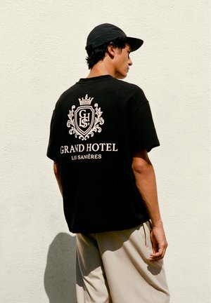 Jonge man met een zwarte pet en een zwart "Grand Hotel Les Sanières" T-shirt, beige broek, staand tegen een lichte gestructureerde muur.