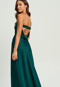 Smaragdgrön satinklänning med en åtsittande bustier, volangrem och öppen rygg. Tyget har en slät, glänsande yta.