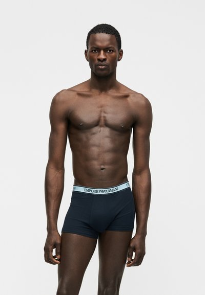Μυώδης άνδρας με σκούρο δέρμα που φοράει μπλε εσώρουχα Emporio Armani, στέκεται μπροστά σε ένα απλό ανοιχτό γκρι φόντο.