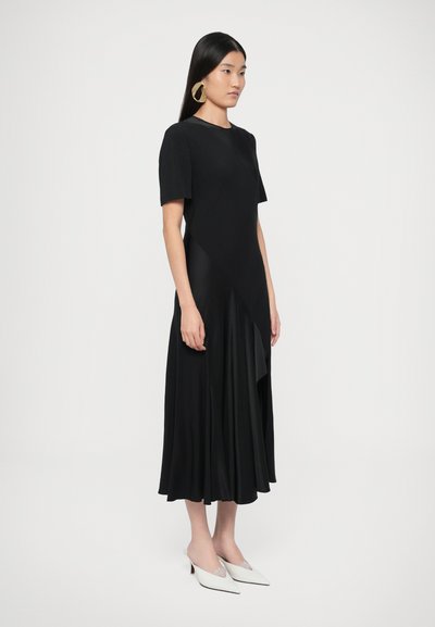 Fekete ruha rövid ujjakkal, aszimmetrikus szegéllyel és selymes anyagból. Fehér, hegyes orrú cipőkkel párosítva. Arany karika fülbevalók.