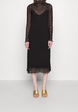 Mujer que lleva un vestido largo negro transparente de manga larga con una combinación de tirantes y tacones mule dorados abiertos en los dedos, de pie contra un fondo liso.