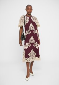 Vestido bordô com aplicações de renda bege. Mangas curtas em renda, na altura do joelho, com padrões intricados. A modelo segura uma pequena bolsa preta.