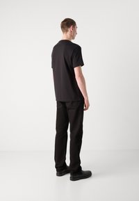 Sort bomuld t-shirt med korte ærmer, rund hals, lagdelt design, parret med sorte bukser og sorte sko, set fra bagsiden.