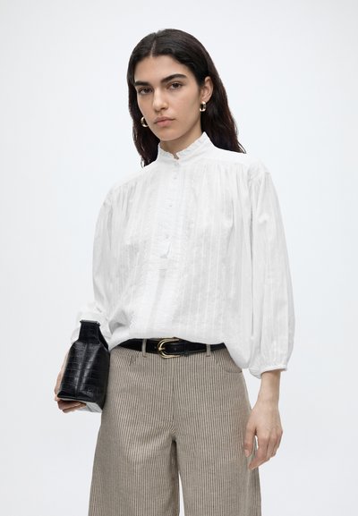 Mujer con cabello largo y oscuro, lleva una blusa blanca abotonada, pantalones a rayas, cinturón negro, pendientes de aro dorados, sosteniendo un bolso negro texturizado.