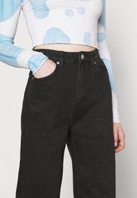 Jeans de mezclilla negros de talle alto con bolsillos frontales, que presentan una textura suave y un diseño clásico de cinco bolsillos. Acompañados de una blusa azul de manga larga con diseño abstracto.