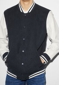 Man med en marinblå och vit varsityjacka med randig krage, manschetter och midjekant, händerna i jackfickorna, blå jeans synliga.