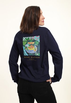 Kvinde iført en navy sweatshirt med Henri Matisses maleri 'The Goldfish' fra 1912 trykt på ryggen, stående med den ene hånd på hoften.