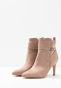 Bottines en suède beige à bout pointu, avec des accents perforés décoratifs et un fin talon aiguille. Elles présentent un détail de boucle sur la sangle.