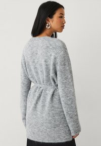 Cardigan grigio a maglia con cintura e finitura morbida e testurizzata; presenta una vestibilità comoda e maniche lunghe. La vista posteriore mostra il dettaglio della vita arricciata.