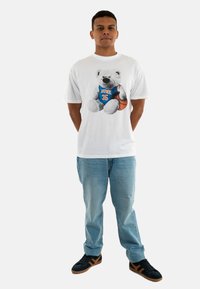 Camiseta de algodón blanca con un gráfico de un oso en una camiseta de baloncesto azul con el texto "DUNK" y el número "35". Combinada con jeans de color azul claro y zapatillas negras.