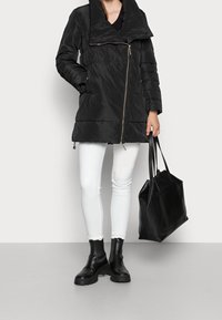 Schwarze Pufferjacke mit großem, umgeschlagenem Kragen und diagonalen Reißverschluss. Getragen über weißen Jeans und kombiniert mit schwarzen Stiefeletten und einer großen Tasche.