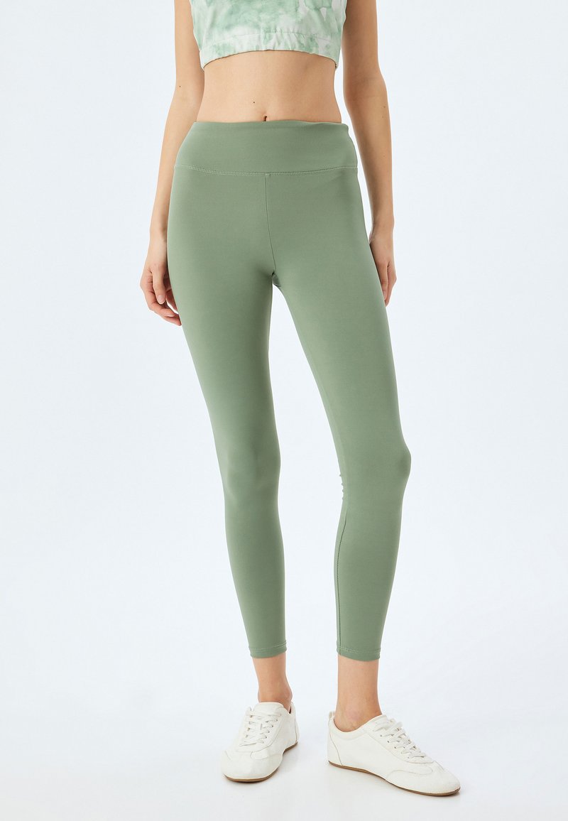 Groene, aansluitende leggings van rekbaar materiaal met een hoge tailleband. Dikke, gladde textuur en minimale stiksels. Witte sneakers gedragen.