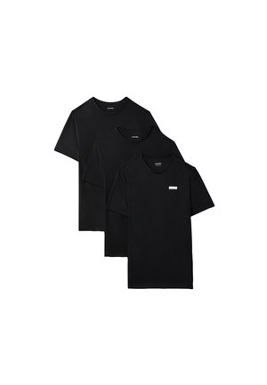 Jeune homme debout, pieds nus, portant un t-shirt noir à manches courtes et un pantalon de jogging noir sur un fond gris uni.