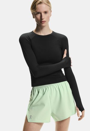 Lys person iført en tætsiddende sort langærmet top og lysegrønne atletikshorts, stående med den ene arm krydset over brystet.