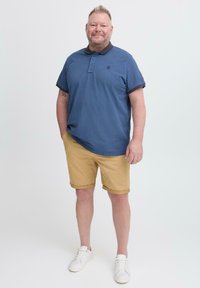 Modra polo majica s temnim ovratnikom, peščene kratke hlače in bele superge. Polo ima logo in zvite rokave. Ohlapen outfit primeren za toplo vreme.