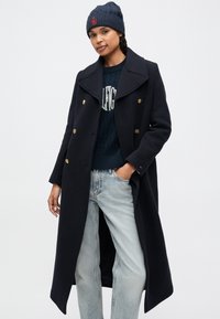 Marineblå uld peacoat med dobbeltradede gyldne knapper, store kraver og sidelommer, båret over en marineblå striktrøje og lyseblå jeans.