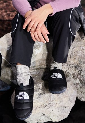 Personne assise sur un rocher portant des chaussons noirs The North Face, des chaussettes grises, un pantalon noir et un haut violet à manches longues, les mains reposant sur les genoux.
