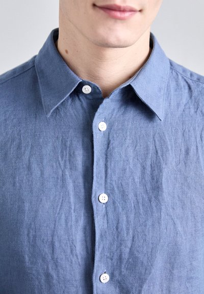 Camisa de lino azul claro con cuello abotonado y cinco botones blancos en el frente. La tela tiene una apariencia texturizada y ligeramente arrugada.