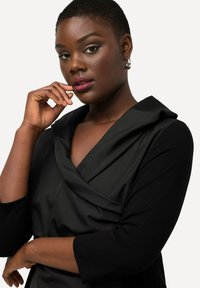 Robe noire avec un décolleté drapé et des manches trois-quarts, dotée d'un tissu lisse et d'un design élégant et structuré.