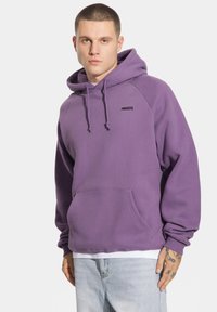 Fioletowa bluza z kapturem, z przednią kieszenią, sznurkiem do regulacji kaptura i małym czarnym logo. Charakteryzuje się luźnym krojem i ściągaczami na mankietach. Nosi się ją na białą koszulę.