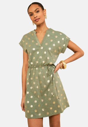 Femme portant une robe courte olive à manches courtes avec des motifs dorés en forme de rayons de soleil, col en V, taille nouée, bracelets en or et boucles d'oreilles sur un fond uni.