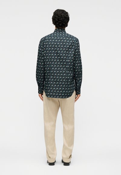 Άνδρας με σγουρά σκούρα μαλλιά στέκεται με την πλάτη, φορώντας μαύρο πουκάμισο με σχέδια μακρυμάνικο και μπεζ παντελόνι, μπροστά σε απλό φόντο.