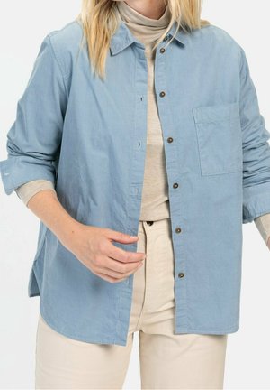 Osoba ubrana w błękitną sztruksową koszulę zapinaną na guziki, na beżowym golfie i beżowych spodniach, z rękawami swobodnie podwiniętymi.