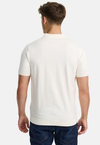 Polo de manga corta en tejido de punto crema con cuello y puños acanalados, que presenta un diseño sin costuras y una textura suave. Combinado con unos vaqueros oscuros.
