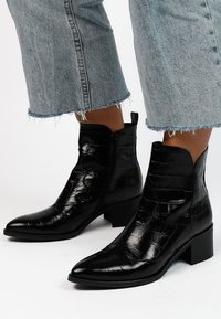 Schwarze Knöchelstiefel aus glänzendem, krokodilgeprägtem Leder, mit spitzer Zehe, seitlichem Reißverschluss und klobigem Absatz.
