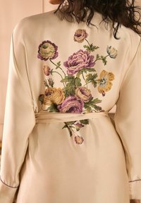 Robe floreale in satin crema con ricami floreali viola, gialli e verdi sulla schiena; presenta maniche lunghe e una cintura da legare.