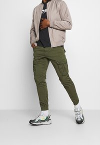 Veste bombers marron clair, t-shirt graphique noir, pantalon cargo olive avec poches latérales, et chaussures de sport aux couleurs mélangées.