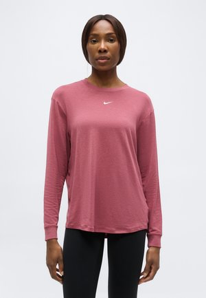 Femme debout portant un t-shirt rose à manches longues Nike avec un logo blanc et des leggings noirs sur un fond blanc uni.