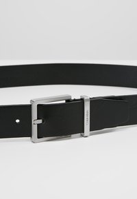 Cintura in pelle nera con una fibbia rettangolare in metallo argentato, caratterizzata da un logo sottile e una finitura liscia per un aspetto pulito.