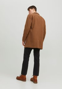 Cappotto di lana marrone con un design strutturato, chiusura frontale a un solo petto e tasche laterali, abbinato a pantaloni neri e stivali di pelle marrone.