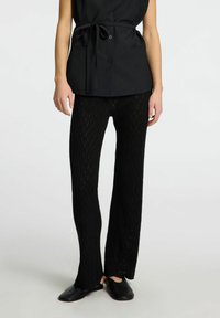 Pantalones de punto negros con un patrón de diamante texturizado, cintura ajustada y piernas acampanadas, combinados con zapatos negros sin cordones.