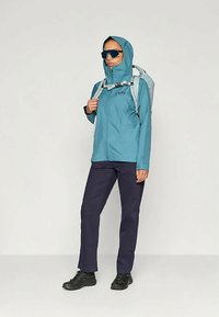 Teal vanntett jakke med hette, kombinert med marineblå bukser og svarte sko. Modellen bærer en lysegrå ryggsekk og har på seg mørke solbriller.