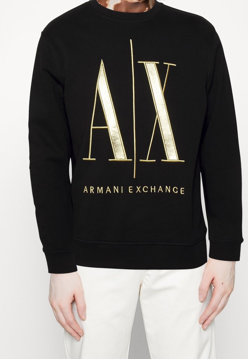 Personne portant un sweat-shirt noir avec de grands textes dorés "A | X" et "Armani Exchange", associé à un pantalon blanc sur fond blanc.