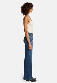 Top tank beige chiaro e jeans svasati blu, con vita alta, tasche e cuciture a contrasto. Il modello è in piedi di lato su uno sfondo semplice.