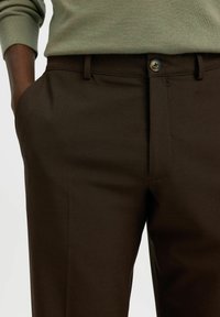 Personne portant un pantalon sur mesure marron foncé avec une fermeture à bouton et zip, main dans la poche, et un haut à manches longues côtelé vert clair.