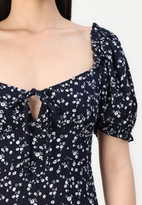 Vestido floral azul-marinho com mangas bufantes, apresentando pequenas flores brancas, um decote quadrado e um detalhe de laço no busto. Tecido texturizado.