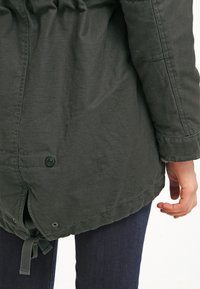 Giacca verde, leggermente strutturata, con cordoncino in vita, dettagli in bottone e orlo arrotondato. Design casual con maniche lunghe.